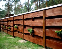 Wooden fence panels with horizontal slats and potted plants attached, backed by evergreen trees